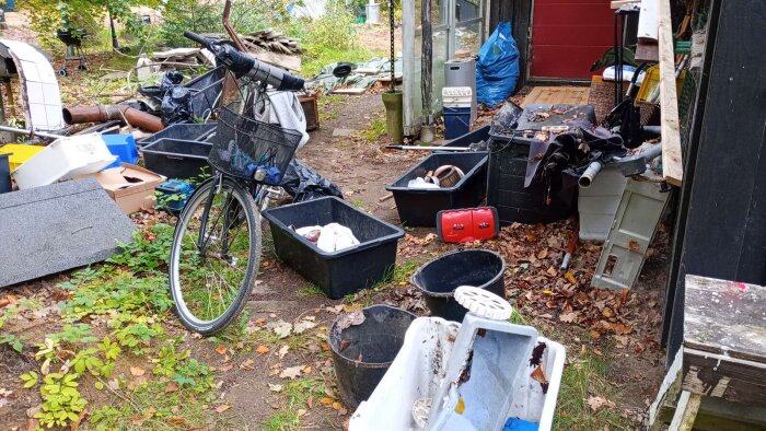 Cykel och flera svarta förvaringslådor omgivna av diverse bråte och trädgårdsföremål, del av en rörig trädgård.