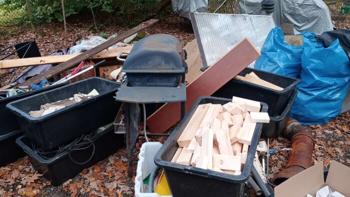 Svart plastlåda med träbitar, grill och diverse föremål utomhus. Träd och löv i bakgrunden.