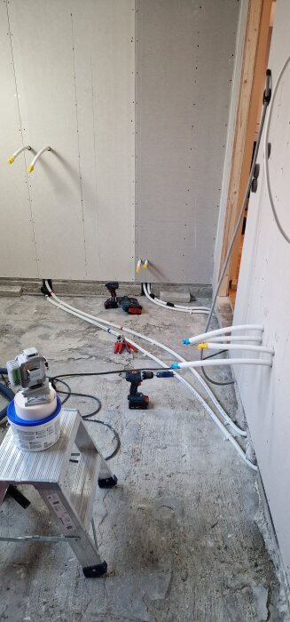Elektrisk borr med verktyg placerade på ett betonggolv. Vita rör är dragna i väggen för VVS-installation.
