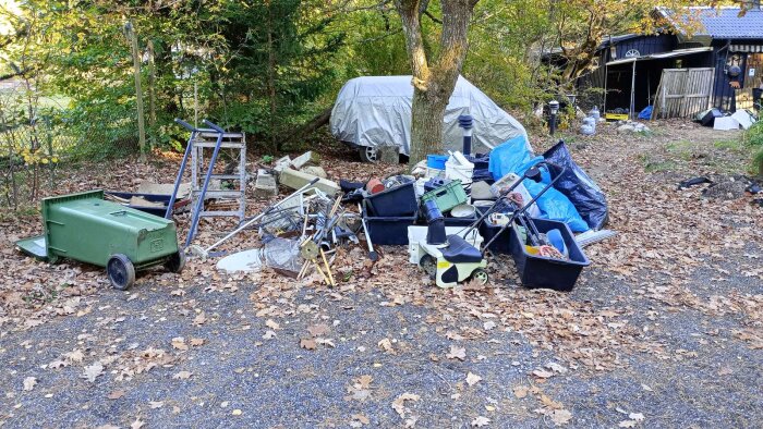 En hög med byggskräp och gamla föremål på marken vid sidan av ett hus, omgiven av träd och löv, med en grön soptunna liggande omkull.