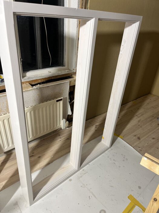 Målat fönsterkarm av trä i en inomhusmiljö, står på golvet framför ett element och en gammal fönsteröppning utan glas.