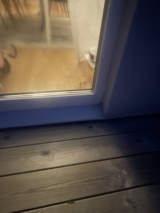 Vy genom glasdörr där ett trägolv syns utomhus och ett inomhus med stolar och bord i bakgrunden.