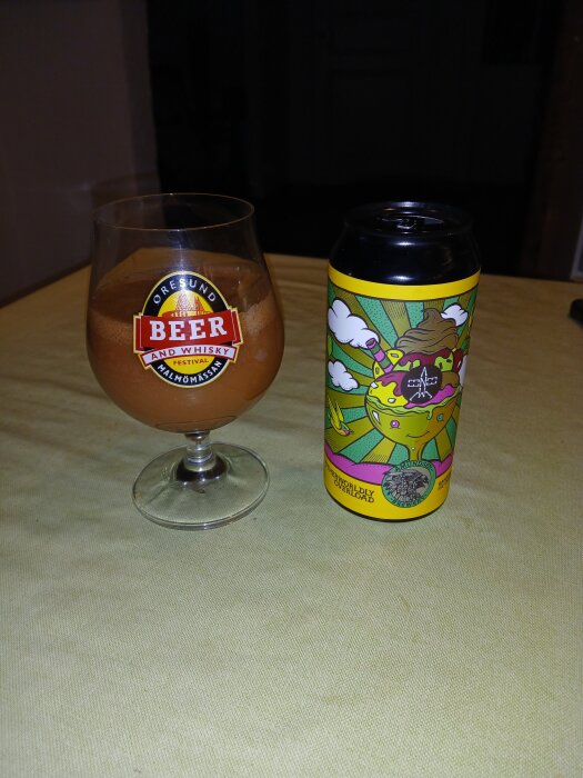 Ett ölglas med en ljusbrun dryck och en färgglad burk med öletikettdesign på ett bord.