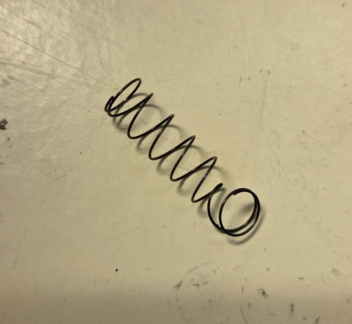 En metallfjäder ligger på ett ljust underlag, troligen ursprungligen från en Makita SP6000J sänksåg.