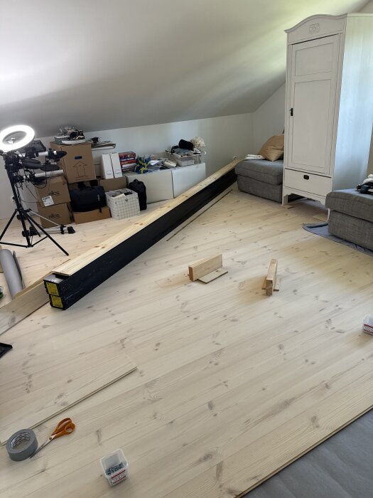 Ett rum under renovering med oordning, innehållande träplankor, verktyg, ett vitt skåp och en soffa.