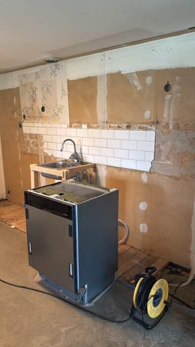 Kök under renovering med en demonterad diskmaskin, halvfärdiga väggplattor och ett provisoriskt monterat handfat.