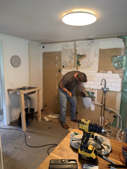Man arbetar med renovering av ett kök, använder verktyg för att ta bort gammal tapet från väggen; skruvdragare och verktyg i förgrunden.