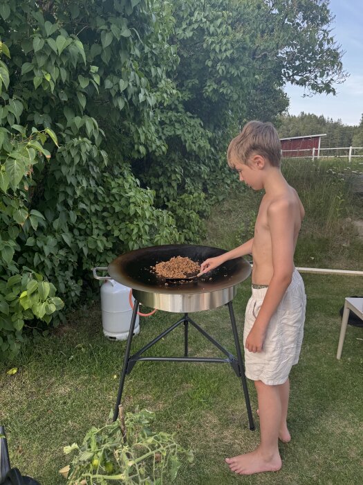 Pojke i shorts lagar mat på en utomhusgrill i trädgården.