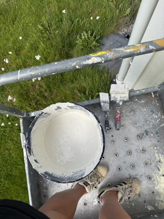 Person målar fasad med vit färg från hink, pensel och roller syns, stående på byggställning, gräs i bakgrunden.
