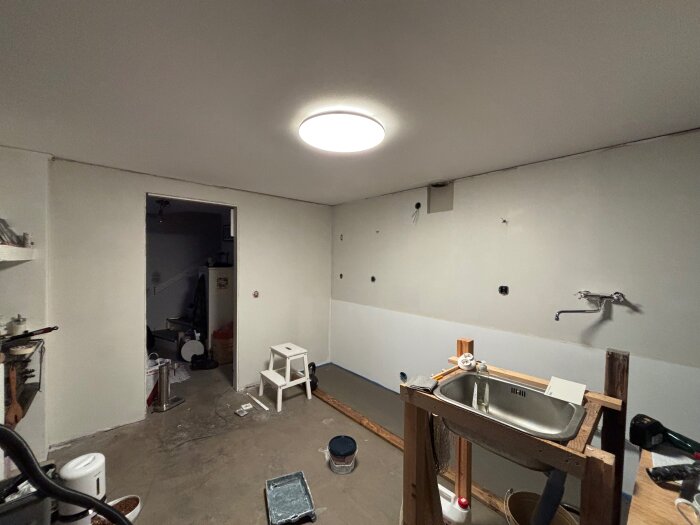 Renovering av ett kök med omålat golv, en provisorisk diskbänk och väggmonterad kran. Extra verktyg och material syns också i rummet.