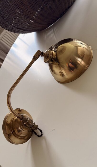 Brun mässingsvägglampa i glänsande metall på vit yta, med spegelbild av person i lampan.