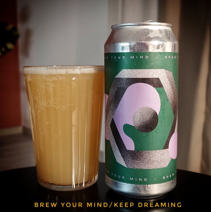 Ett glas ljus öl bredvid en burk med etiketten "Brew Your Mind/Keep Dreaming", mot en suddig bakgrund.