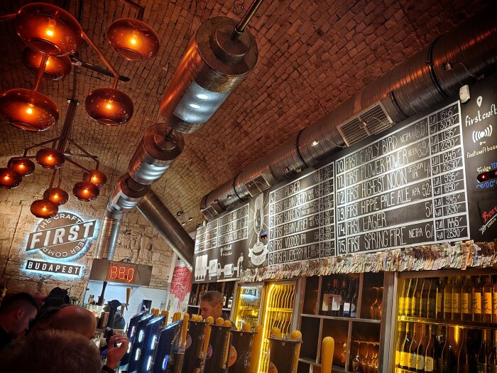 Interiör av ett bryggeri med tegelväggar, upplysta lampor och en meny med olika ölsorter. Neonljus visar "FIRST Craft Beer Budapest".