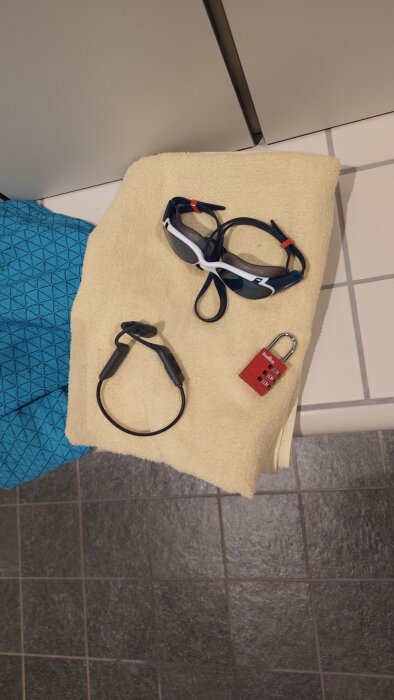 Handduk med simglasögon, hörlurar och hänglås på klinkergolv i omklädningsrum.