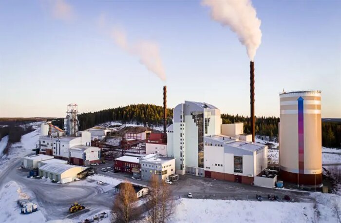Fjärrvärmeverk i Skellefteå i vinterlandskap, med höga skorstenar och rök. Stora byggnader och flisdepåer syns i bakgrunden.