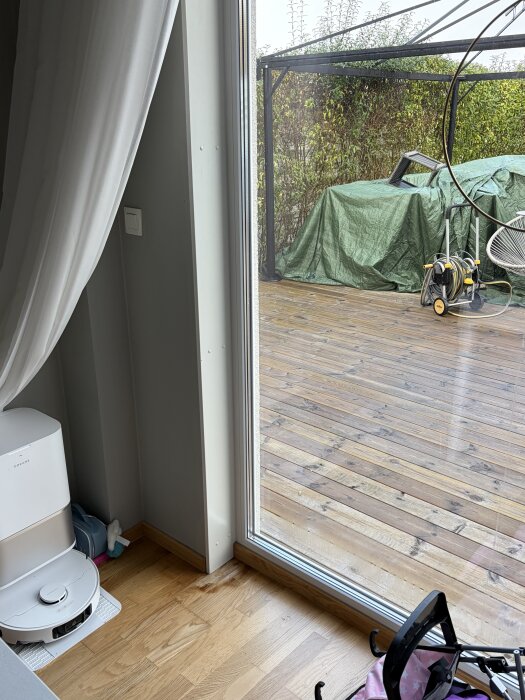 Vy genom glasdörr mot träaltan med grön presenning, växter och trädgårdsredskap. Inomhus syns en robotdammsugare.