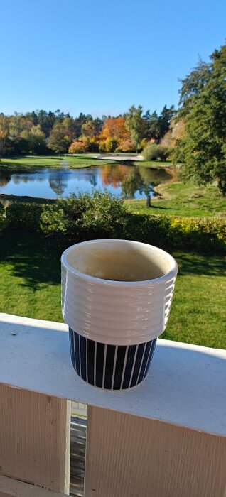 Kaffekopp med macchiato på en altanräcke, med utsikt över en grönskande park och sjö i bakgrunden under en solig dag.