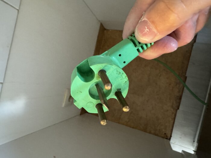 Grön stickkontakt med tre stift hålls i en hand, del av köksmiljö syns i bakgrunden.
