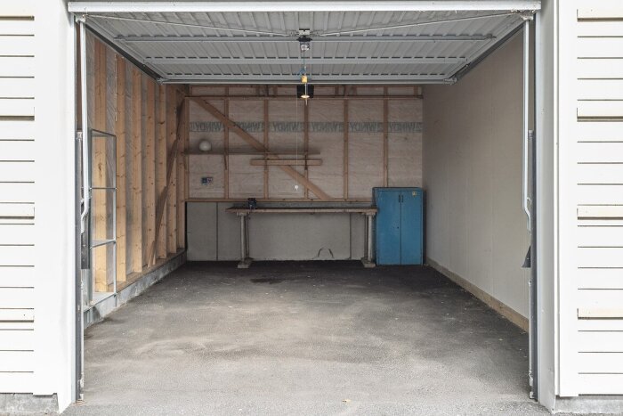 Öppet kallgarage med träpanel och vindduk på väggarna, en arbetsbänk och ett blått skåp, sedd från den öppna garageporten.