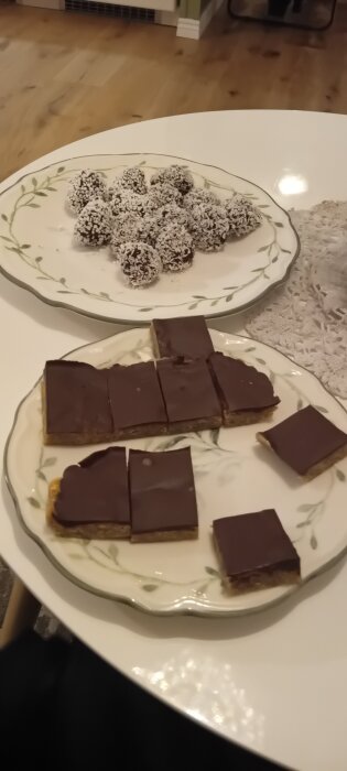 Tallrikar med chokladbollar täckta i kokos och rutor av chokladtoppade jordnötskakor med solrossmör på ett bord.