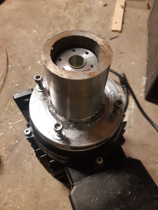 Cylindrisk metalladapter svetsad på bas, monterad på motor för Bergma bergborrslipmaskin. Golv och verktyg synliga i bakgrunden.