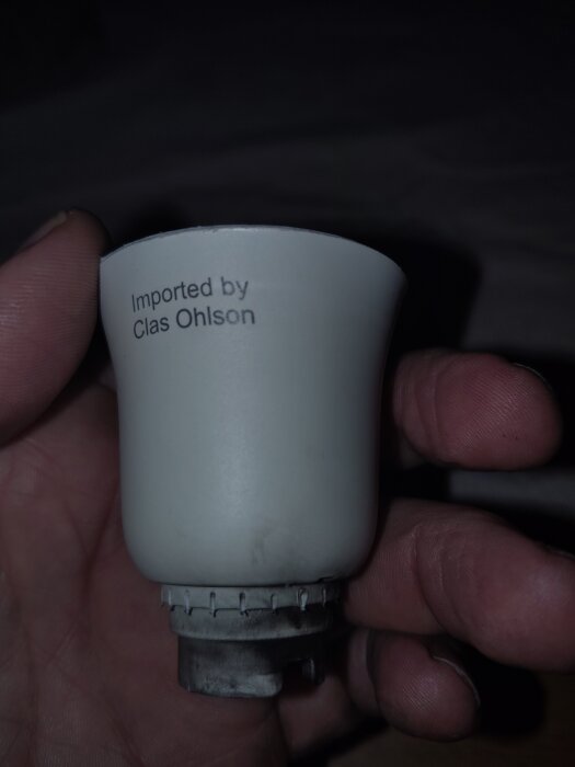 Hand håller en trasig LED-lampa. Text på lampan: "Imported by Clas Ohlson". Burna kanter och skada vid sockeln.