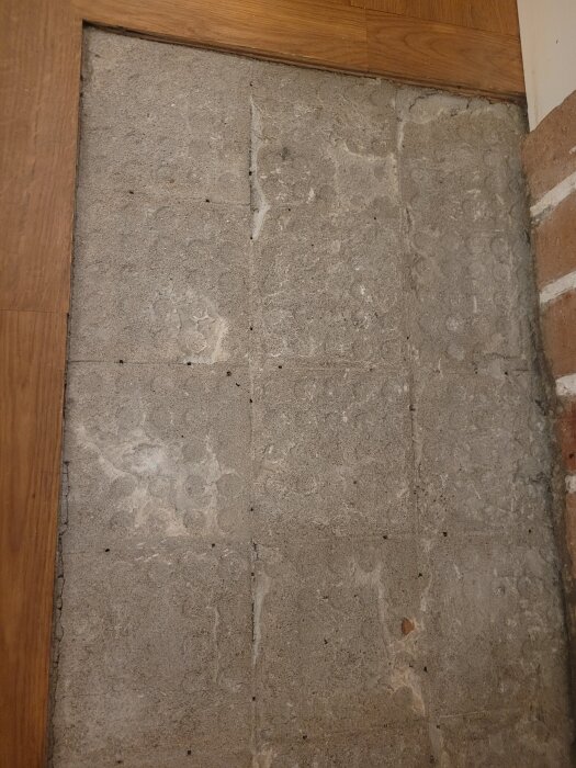 Bilden visar en murad yta med rutor och små hål på betongliknande material, omgivet av träpaneler och en tegelvägg.