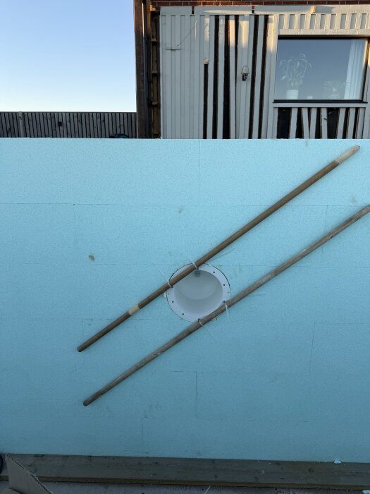 Lampa monterad i blå isoleringsvägg, hålls av två sopskaft och buntband, mot en husvägg.