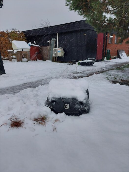 Robotgräsklippare täckt av snö i en vinterträdgård bredvid ett hus och en byggnad, omgiven av snö och frost.