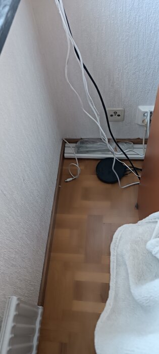 Kablar och en svart rund enhet på golvet intill väggen med eluttag och vit strömlist.