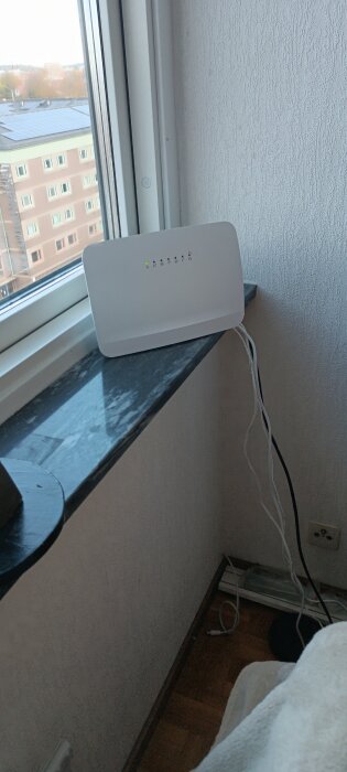 Vit, fyrkantig, digital enhet på fönsterbräda med utsikt över bostadshus, synliga sladdar på sidan.