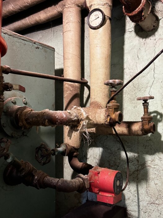 Rörledningar med två tomma kranar och en manometer, omgivna av isolerade rör. En röd pump syns i nedre delen av bilden.