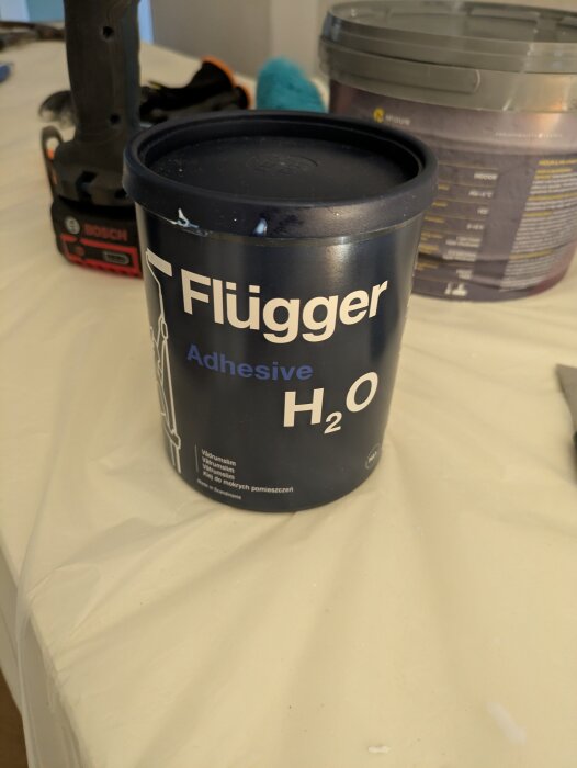 Burk med Flügger H2O vattentät lim på ett bord, med verktyg och andra byggmaterial i bakgrunden.