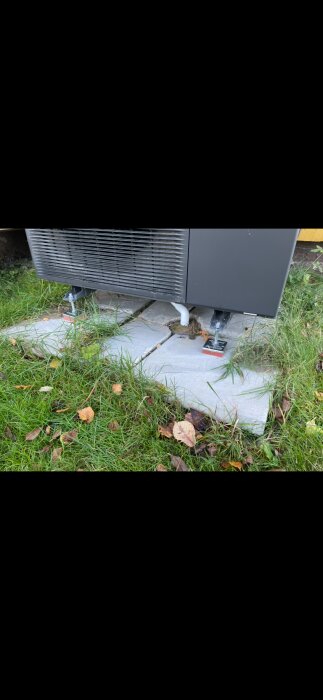 Luft/vatten-värmepump stående på betongplattor som är ojämna; gräs runt om och synliga justeringsfötter.
