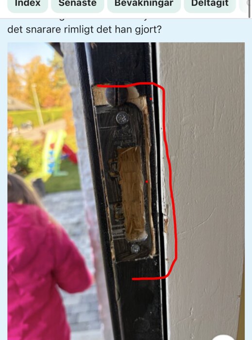 Närbild på en dörrkarm med urtaget trä och synliga skruvar, omgivet av röda markeringslinjer. En person i röd jacka i bakgrunden.