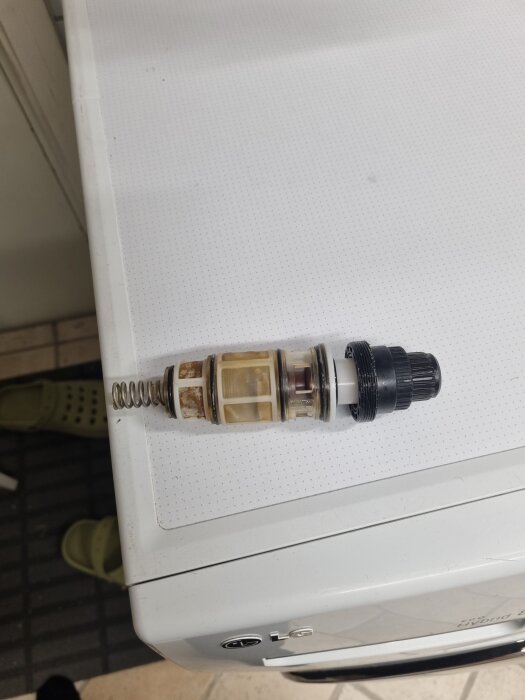 En demonterad del av ett termostatpaket från en blandare ligger på en vit yta, med synlig fjäder och filterdel.
