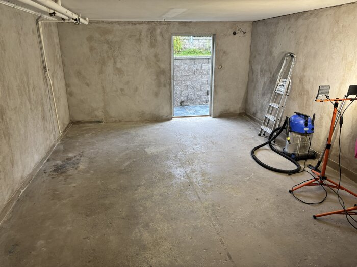 Källare med repigt betonggolv, en stege, dammsugare och belysning; utgång till trädgård synlig genom dörröppning.
