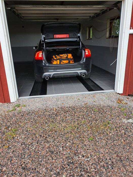 Svart bil parkerad i garage med öppet bagageutrymme, orange väska och två gummimattor på golvet.