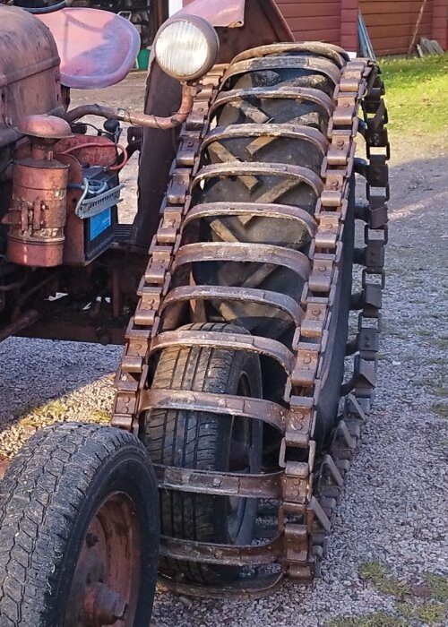 Gammal traktor med stora metallband över däcken, del av front och lampa synliga, står på grus.