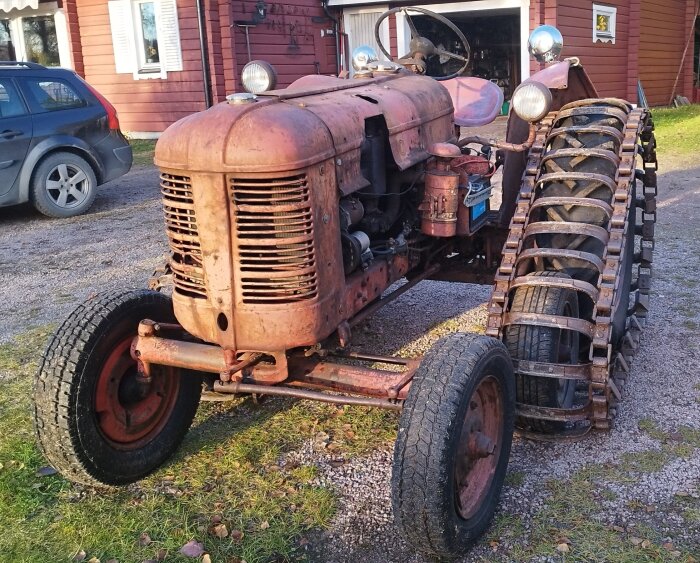 En gammal, rostig traktor med kedjor på bakhjulen står parkerad framför ett hus.