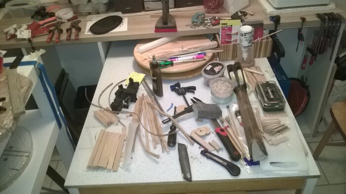 Arbetsyta på köksbord med verktyg, träbitar, penslar och limtuber, omgiven av organiseringstillbehör och diverse redskap.