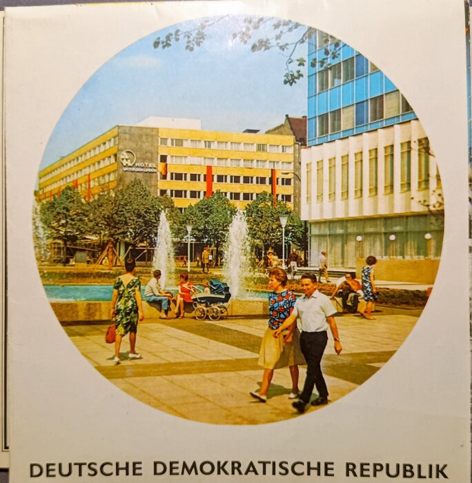 Broschyrbild av ett torg i DDR, med ett hotell, människor som promenerar och fontäner i bakgrunden.