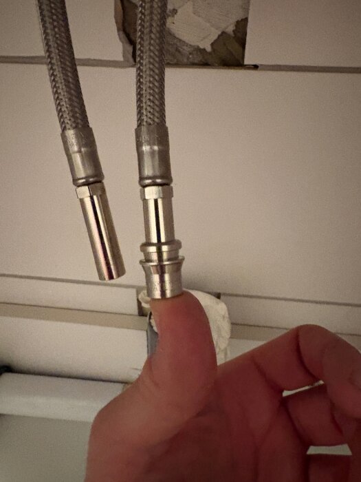 Två flexibla anslutningsslangar för VVS koppling, en hand håller i en slang, med kakel och en vägg i bakgrunden.