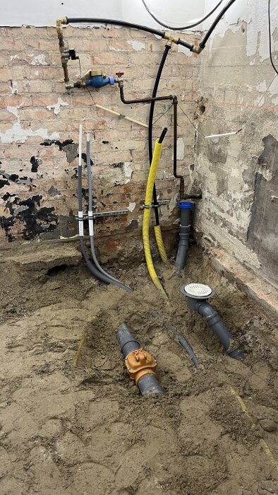 Rörinstallation i byggprojekt med grå och gula rör mot tegelvägg, samt en ventil på marken. Rören är delvis täckta av jord.
