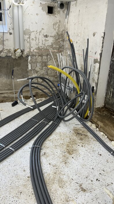 Grå värmerör och gul kabel i en källare under installation av golvvärme med fjärrvärme. Olika rör och kablar ligger på golvet.