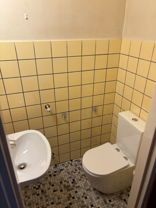 Gäst-wc med gula kakelplattor och ett vitt handfat samt toalett. Väggmonterade rör och hål syns där ett element tidigare suttit.