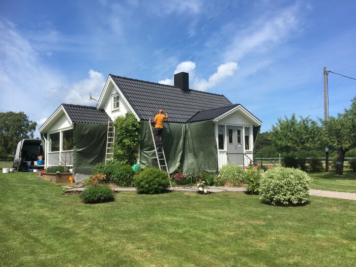 Vit villa med svart tak behandlat med Grön-Fri, person på stege vid huset, grönskande trädgård, klarblå himmel, husbil synlig i bakgrunden.