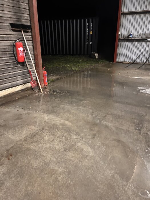 Betonggolv med vattenpöl i ett skjul, omgiven av en spade, en kvast och en brandsläckare vid väggen.