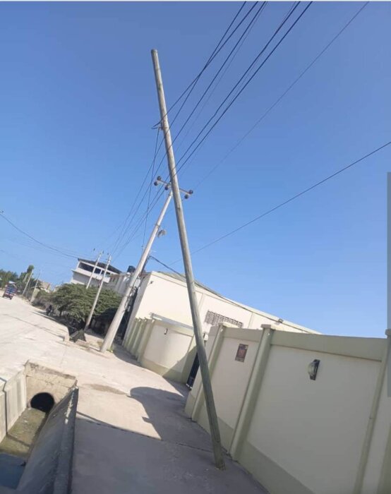 Lutande telefonstolpe nära en väg och byggnader, med elkablar ovanför, mot blå himmel.
