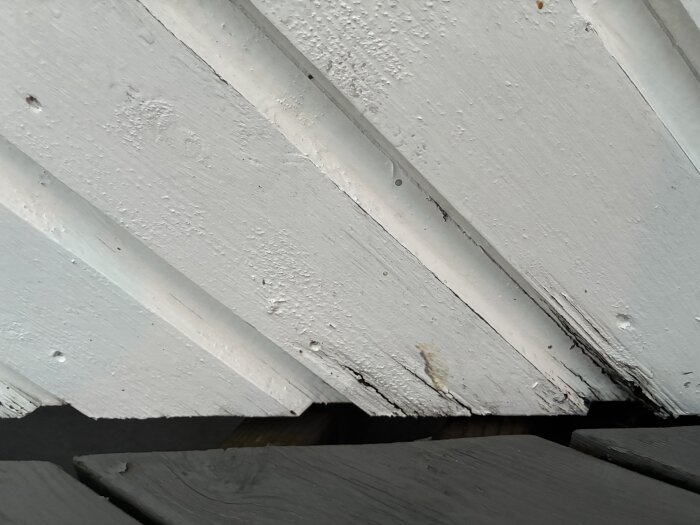 Flagnande vit färg på träpanel utomhus med synliga tecken på slitage och små hål i ytan.